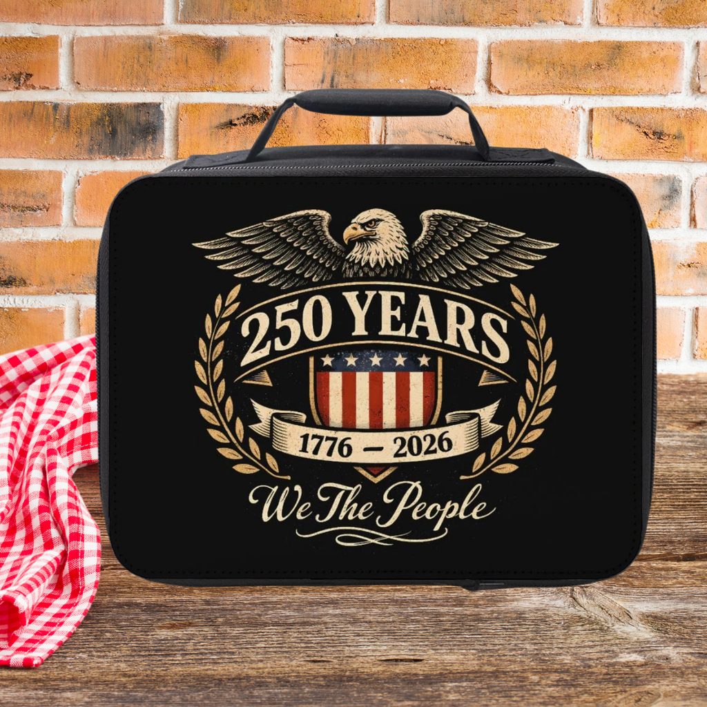 Black lunch bag with eagle design and '250 Years 1776-2026 We The People' text on a wooden picnic table against a brick wall.