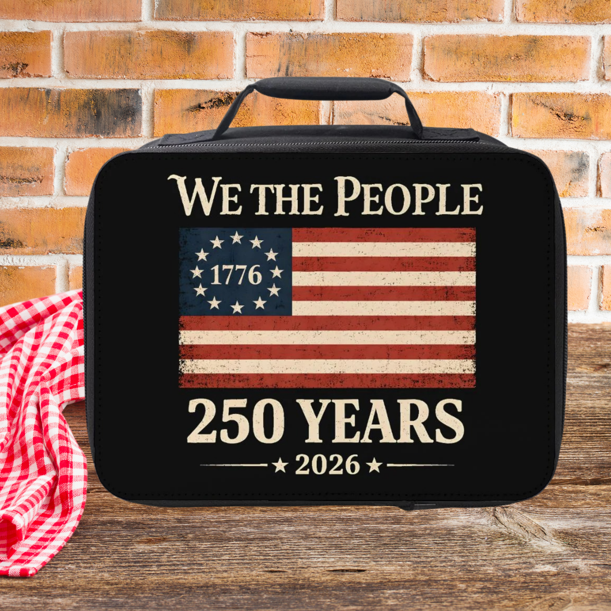 Black bag with American flag design and text 'We the People 1776 250 Years 2026' on a wooden picnic table with a brick wall background.