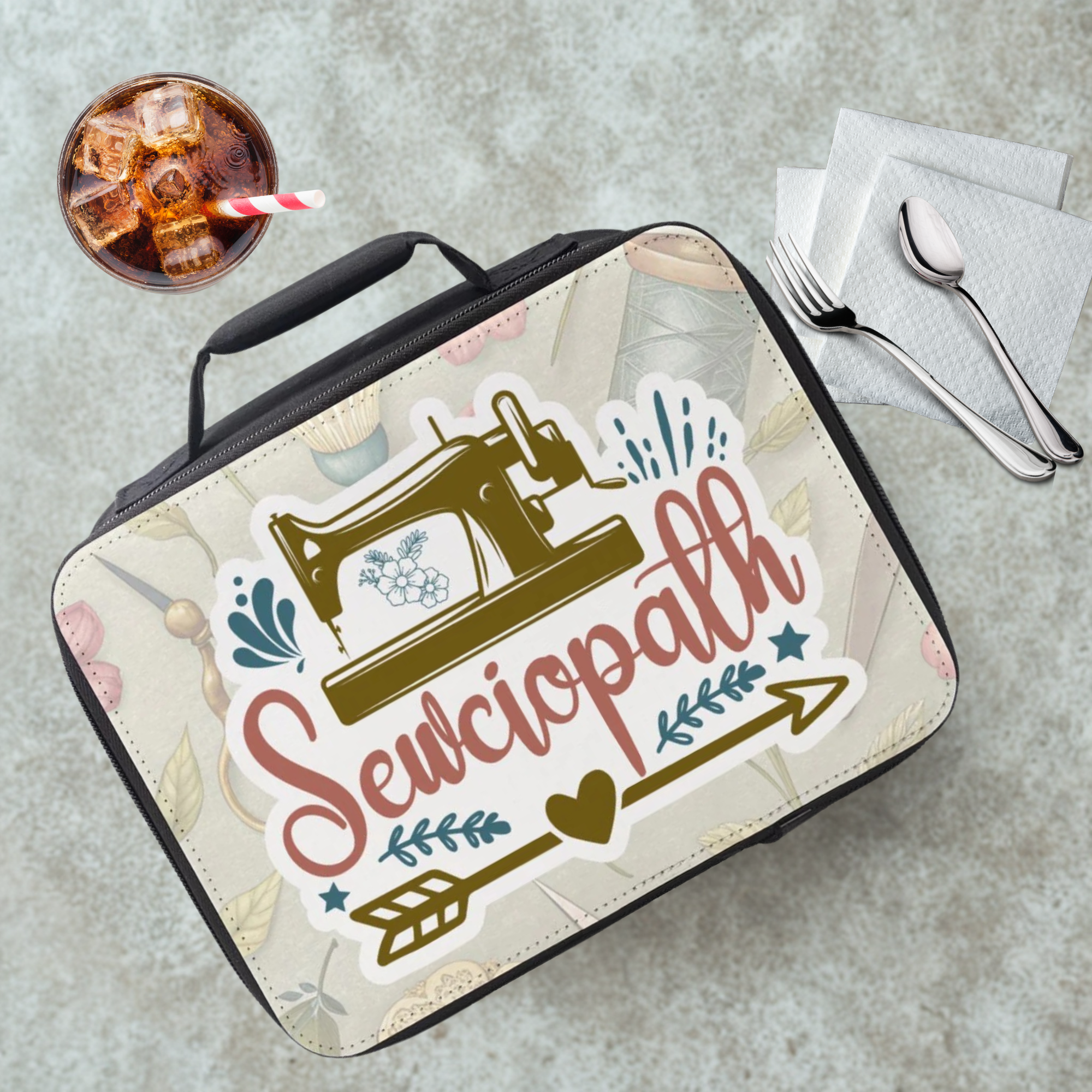 Lunch box with 'Sewciopath' design on a gray cafeteria table with a drink and utensils.