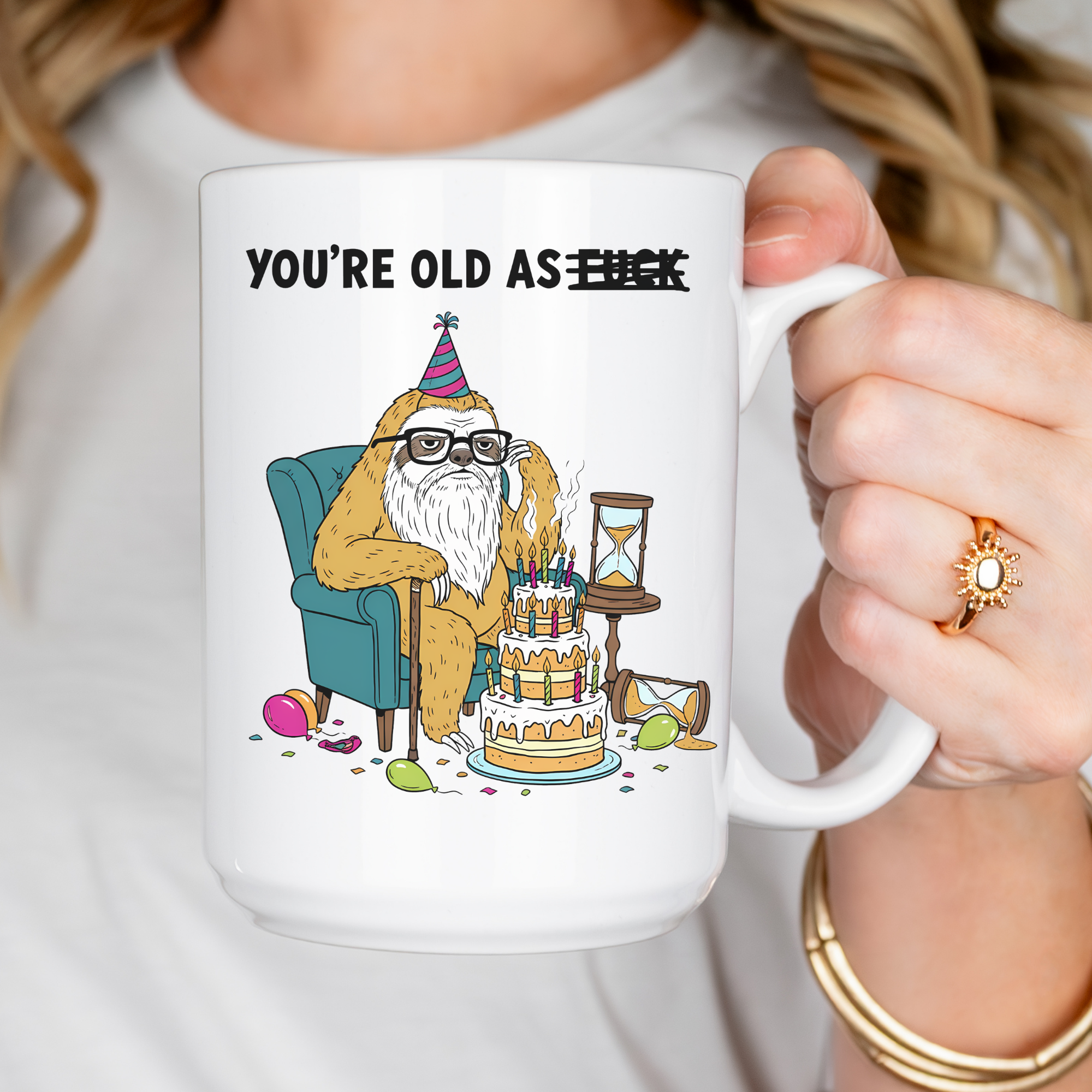 Person holding a mug with a cartoon bear celebrating a birthday and text 'YOU'RE OLD AS', wearing a white shirt.
