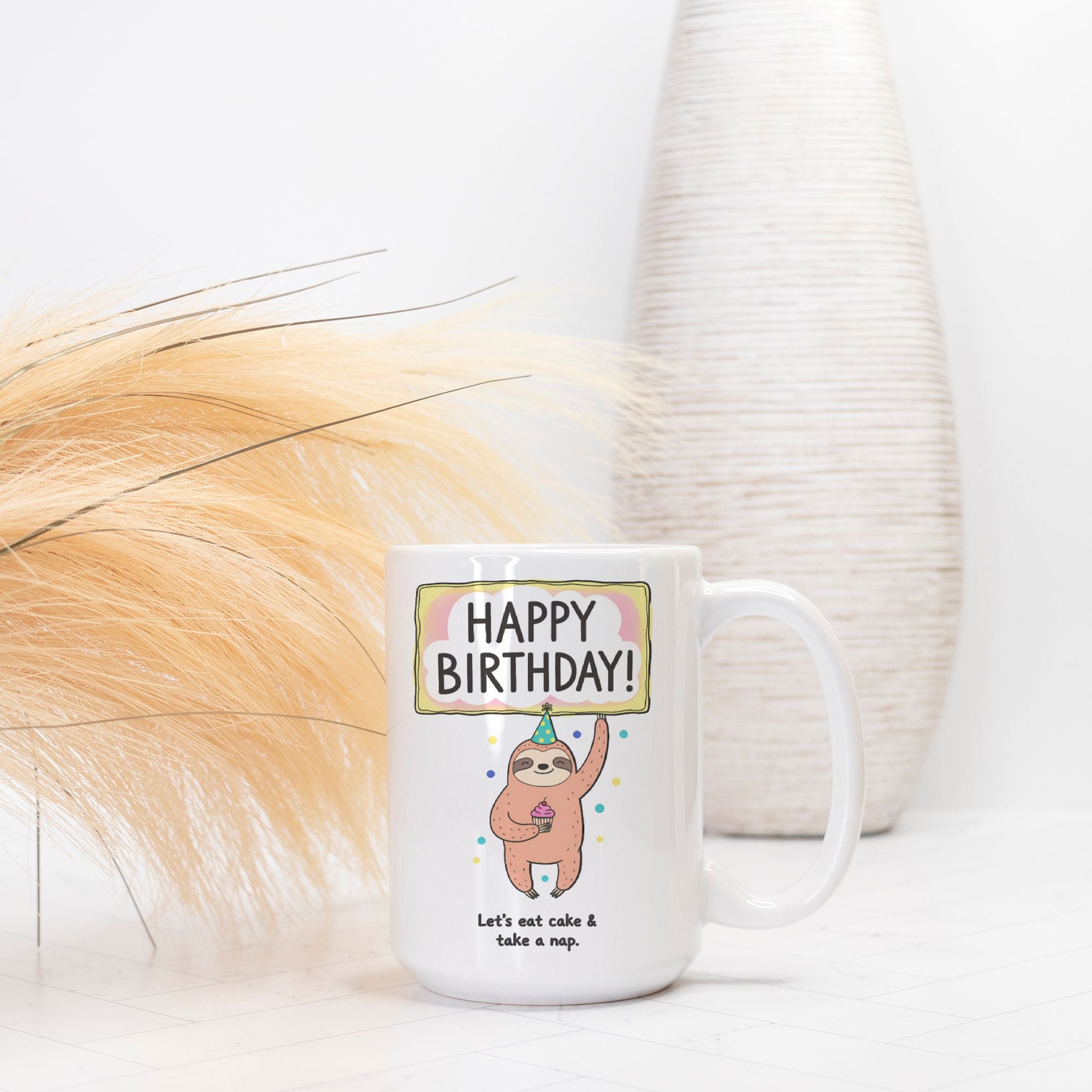 White mug with a cartoon sloth holding a cake and 'Happy Birthday!' text, placed on a light surface with decorative elements.