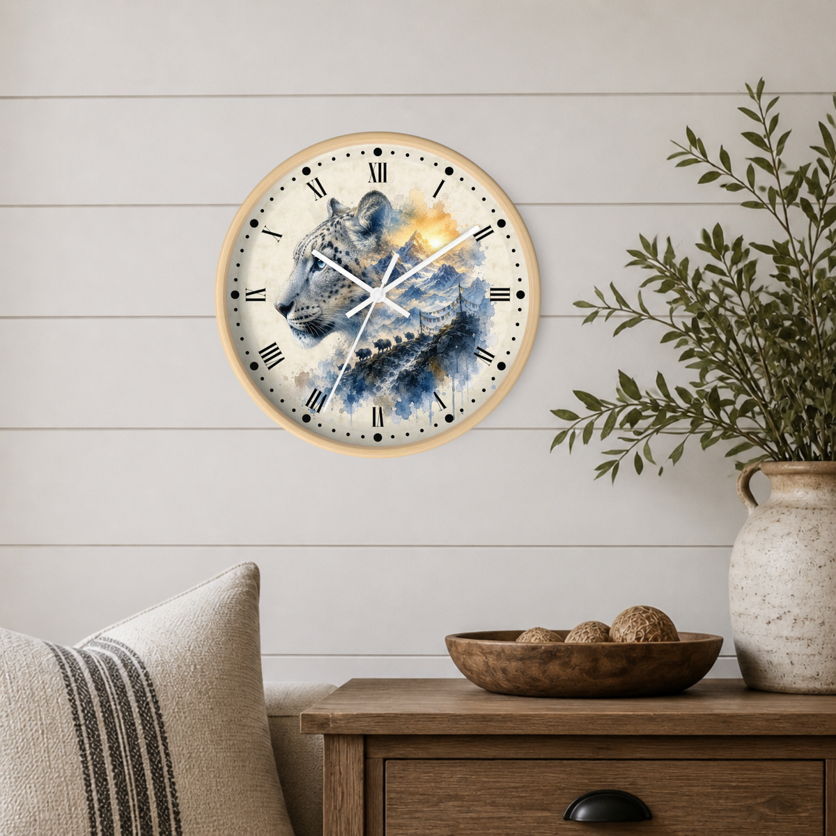 A round wall clock with a light wood frame hangs on a white shiplap wall above a wooden side table. The clock face features a painted snow leopard in blue, gray, and white tones with a warm golden glow.