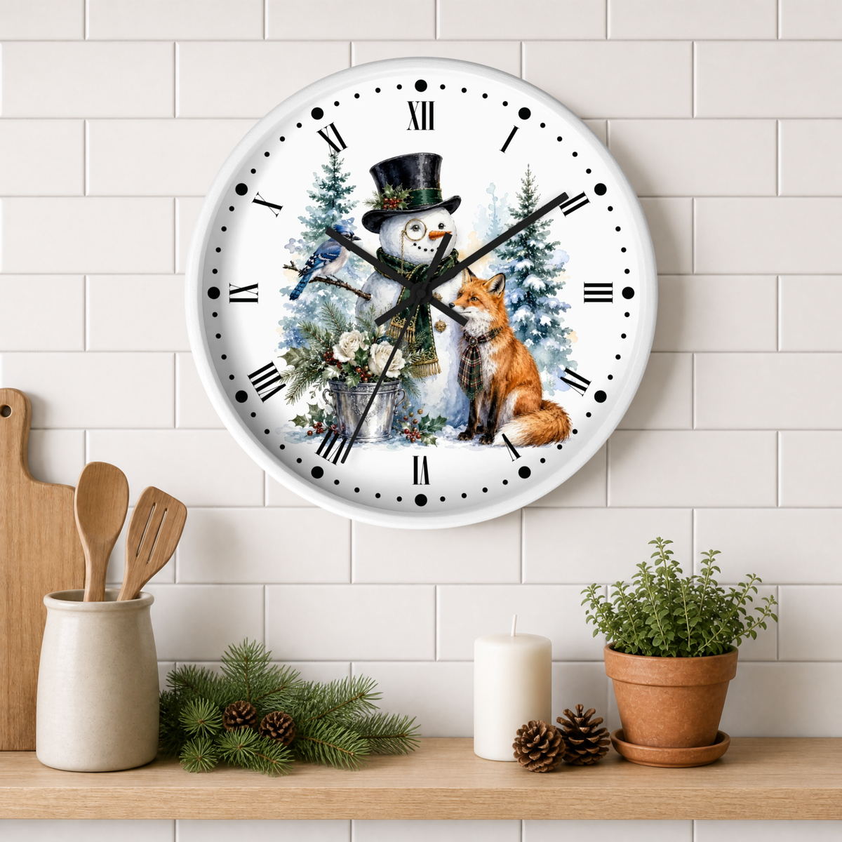 A round wall clock with a white face and black Roman numerals, decorated with a winter scene featuring a snowman in a top hat, a fox, pine trees, and flowers. The clock is mounted on a white subway tile wall above a wooden shelf with kitchen utensils, pine branches, a candle, and a potted plant.