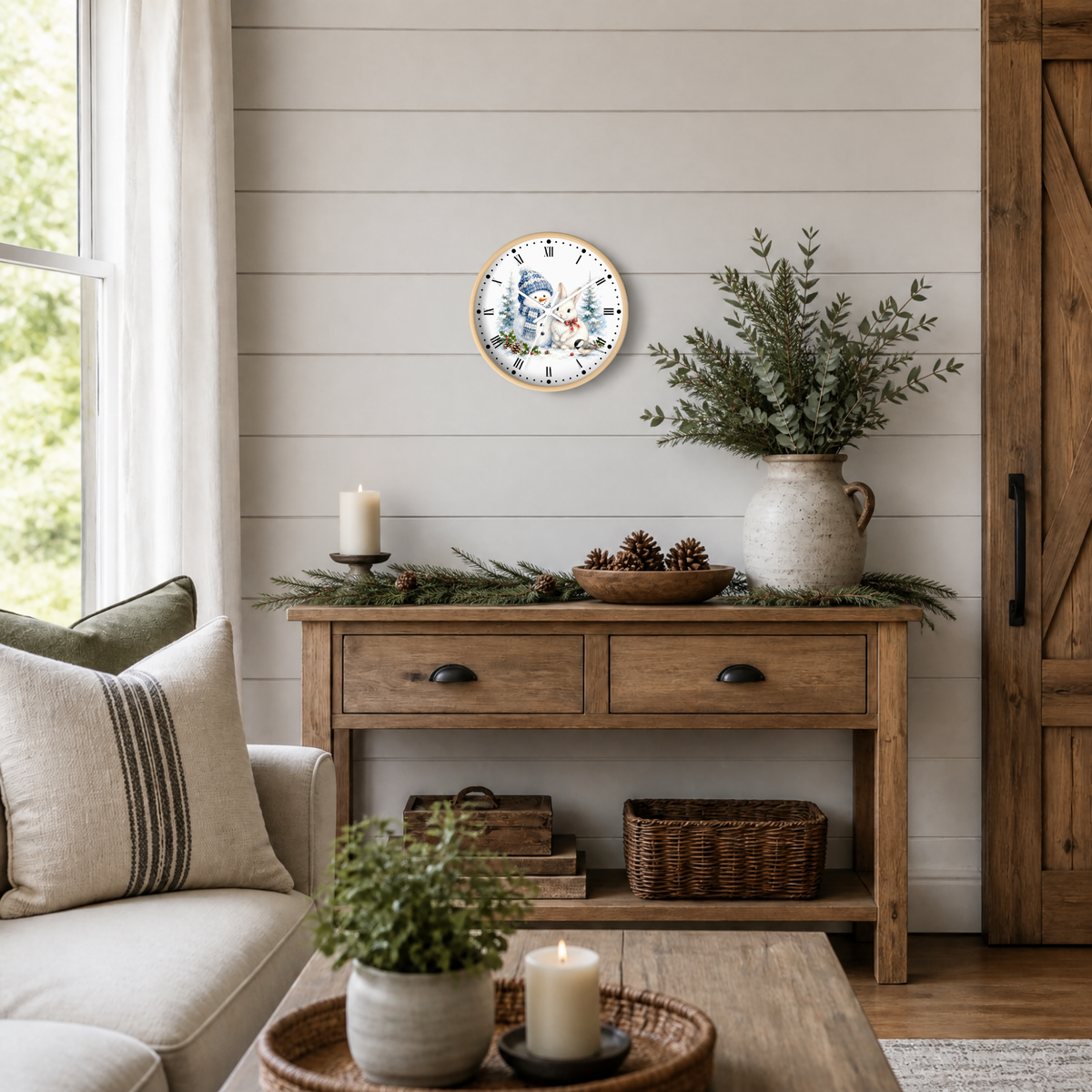 A round wall clock with a blue and white snowman and rabbit illustration and a gold-toned frame hangs on a white shiplap wall above a rustic wooden console table styled with greenery and candles.