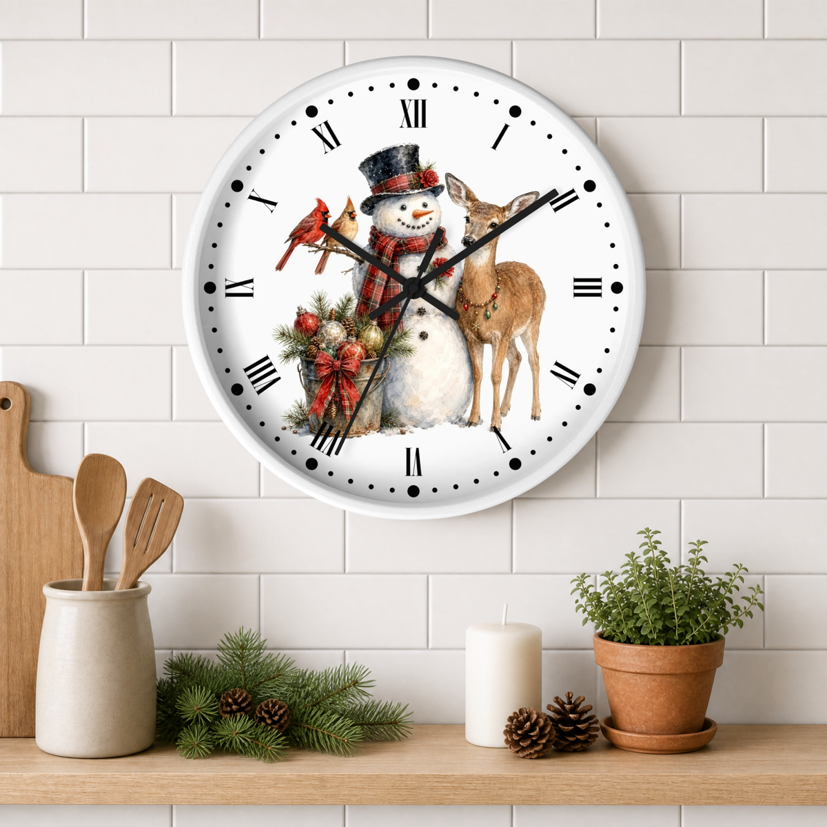 A round wall clock with a white face and black Roman numerals, decorated with a winter holiday illustration of a snowman, a deer, a bird, and red birds. The clock is mounted on a white subway tile wall above a wooden shelf styled with kitchen utensils, pine branches, a candle, and a potted plant.