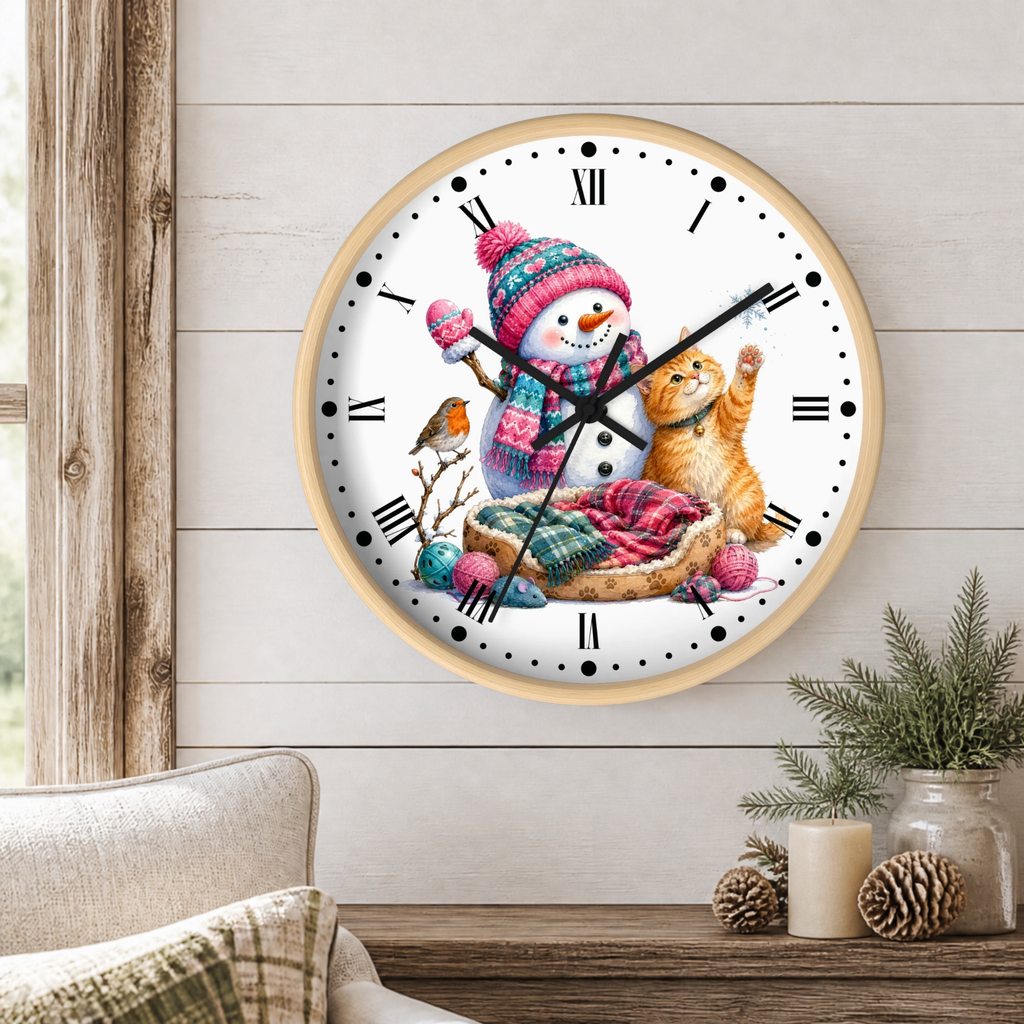 A round wall clock with a light wood frame hangs on a white shiplap wall above a rustic wooden table. The clock face features a winter illustration with a snowman in a pink and blue knit hat and scarf, a ginger cat, a small bird, and a bed of colorful wool.