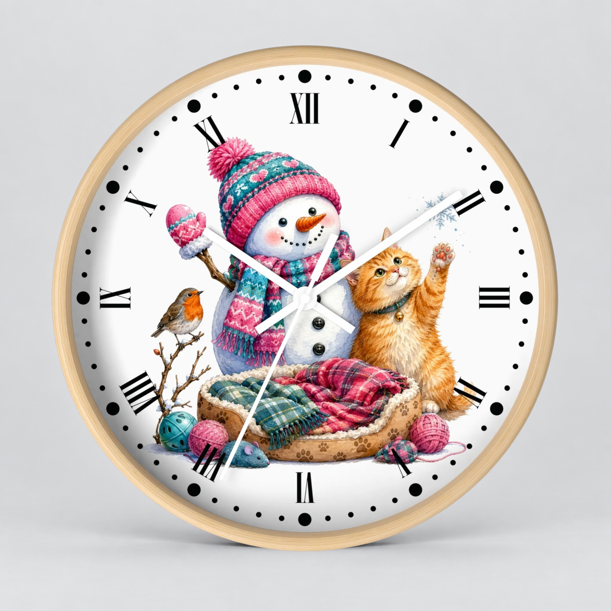 A round wall clock with a light wood frame and a white face, featuring a colorful winter illustration of a snowman in a pink knit hat and scarf, an orange tabby cat, a small bird, and a bed with plaid blankets.