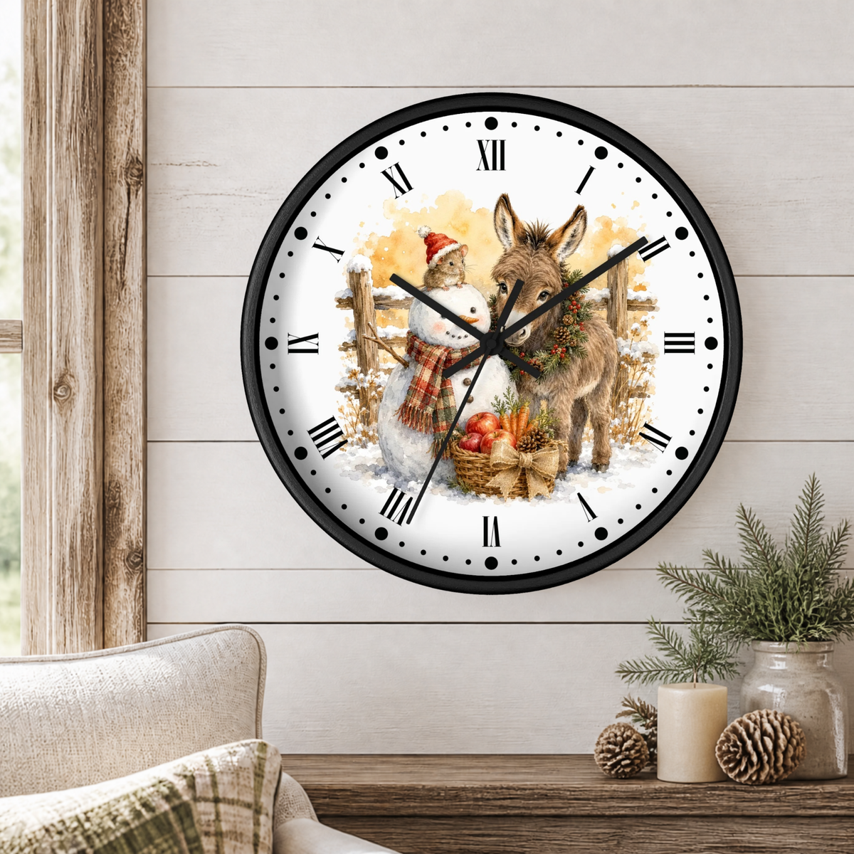 A round wall clock with a black frame and a festive winter illustration of a snowman and a donkey, mounted on a white wood-paneled wall above a wooden table with decorative pine branches and candles.