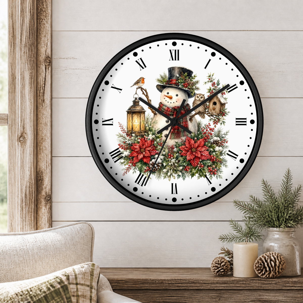 A round wall clock with a black frame and a white face, decorated with a festive Christmas design featuring a snowman, red poinsettias, green foliage, a bird, a lantern, and a birdhouse. The clock is mounted on a light wood-paneled wall above a wooden table with a candle and pinecones.