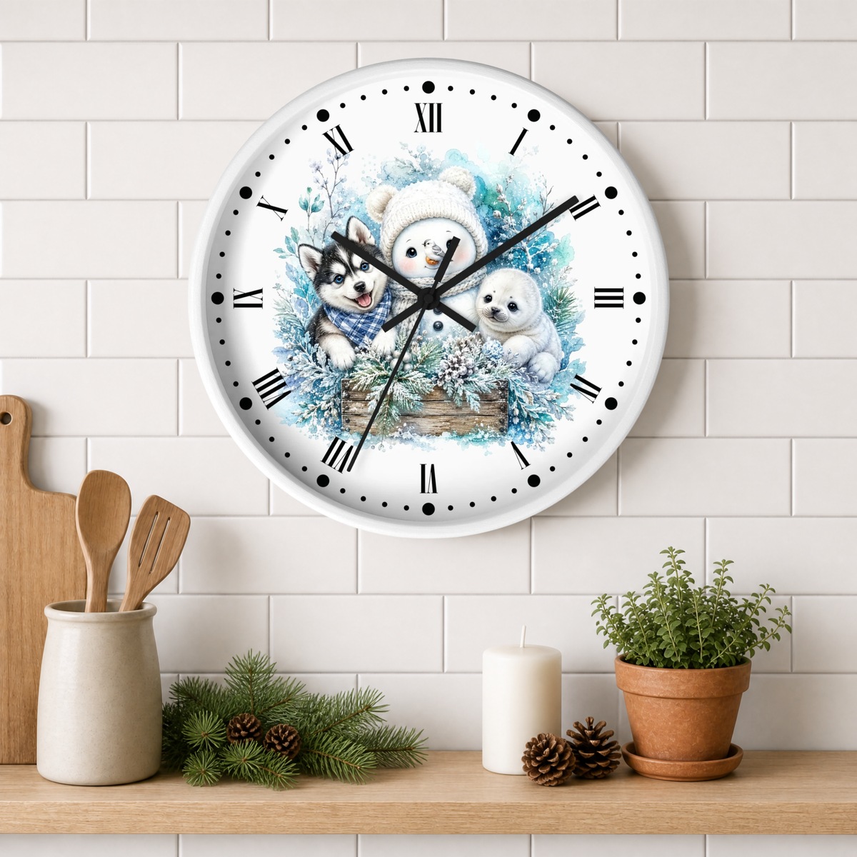 A round wall clock with a white face and black Roman numerals, decorated with an illustration of a snowman, husky, and seal in a basket. The clock is mounted on a white subway tile wall above a wooden shelf styled with kitchen utensils, pine branches, a candle, and a potted plant.