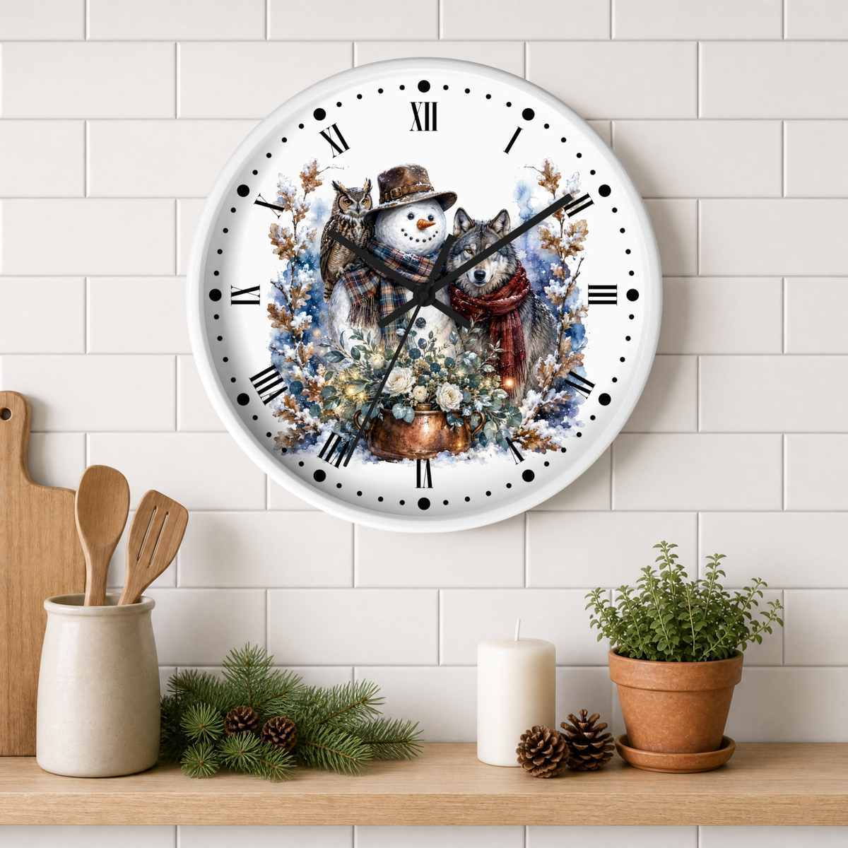 A round wall clock with a white face and black Roman numerals, featuring a winter illustration of a snowman ,a wolf , and owl in a cozy winter scene. The clock is mounted on a white subway tile wall above a wooden shelf styled with kitchen utensils, pine branches, a candle, and a potted plant.