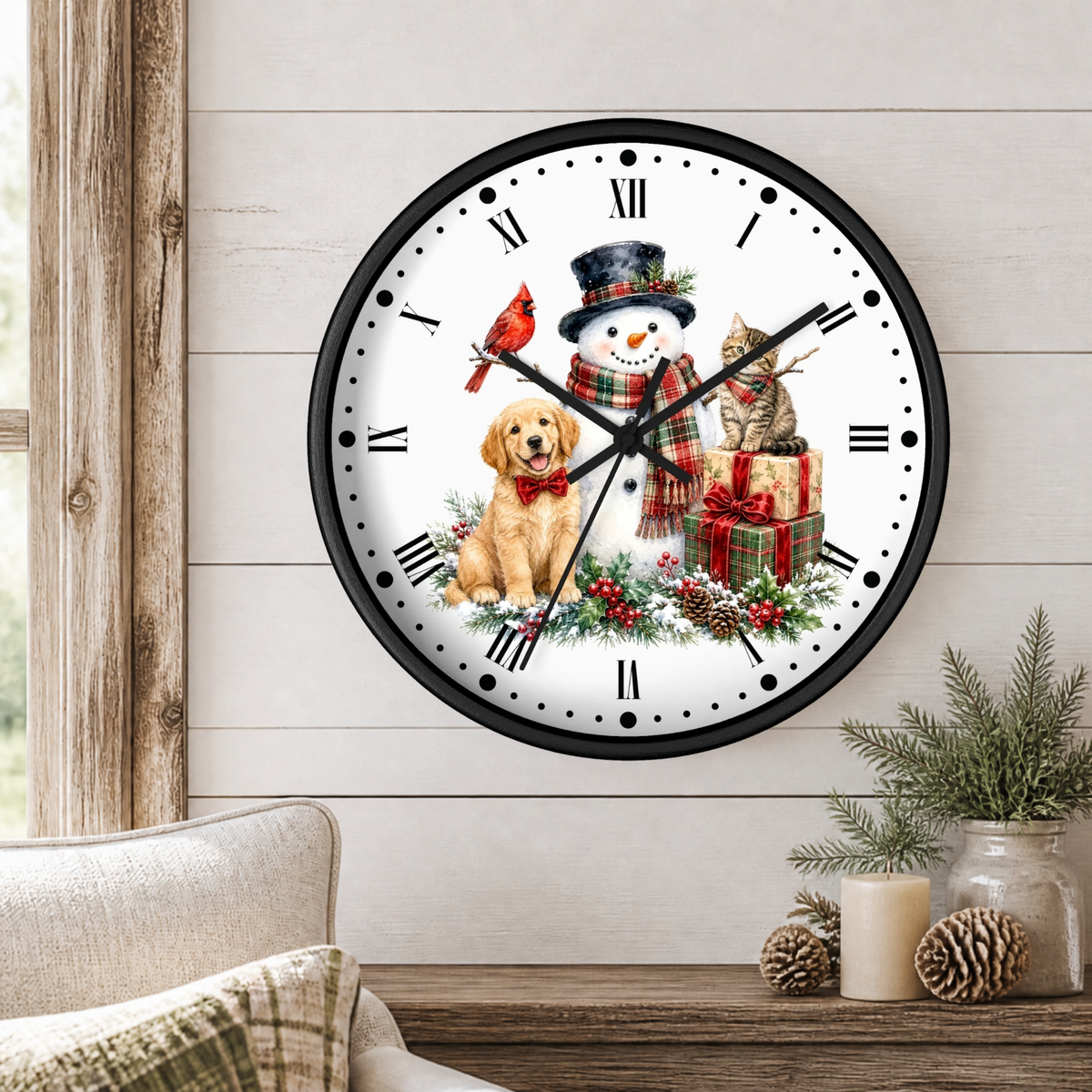 A round wall clock with a black frame and a white face, decorated with a Christmas holiday scene featuring a snowman, a golden retriever dog, a red cardinal, a tabby cat, pinecones, and wrapped gifts. The clock is mounted on a light wood-paneled wall above a rustic wooden table with a candle and pinecones.