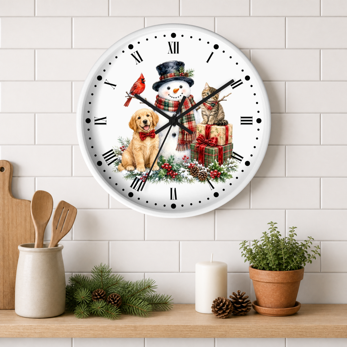 A round wall clock with a white face and black Roman numerals, decorated with a festive Christmas scene featuring a snowman, a golden retriever, a tabby cat, a red cardinal, and wrapped gifts. The clock is mounted on a white subway tile wall above a wooden shelf styled with kitchen utensils, pine branches, a candle, and a potted plant.