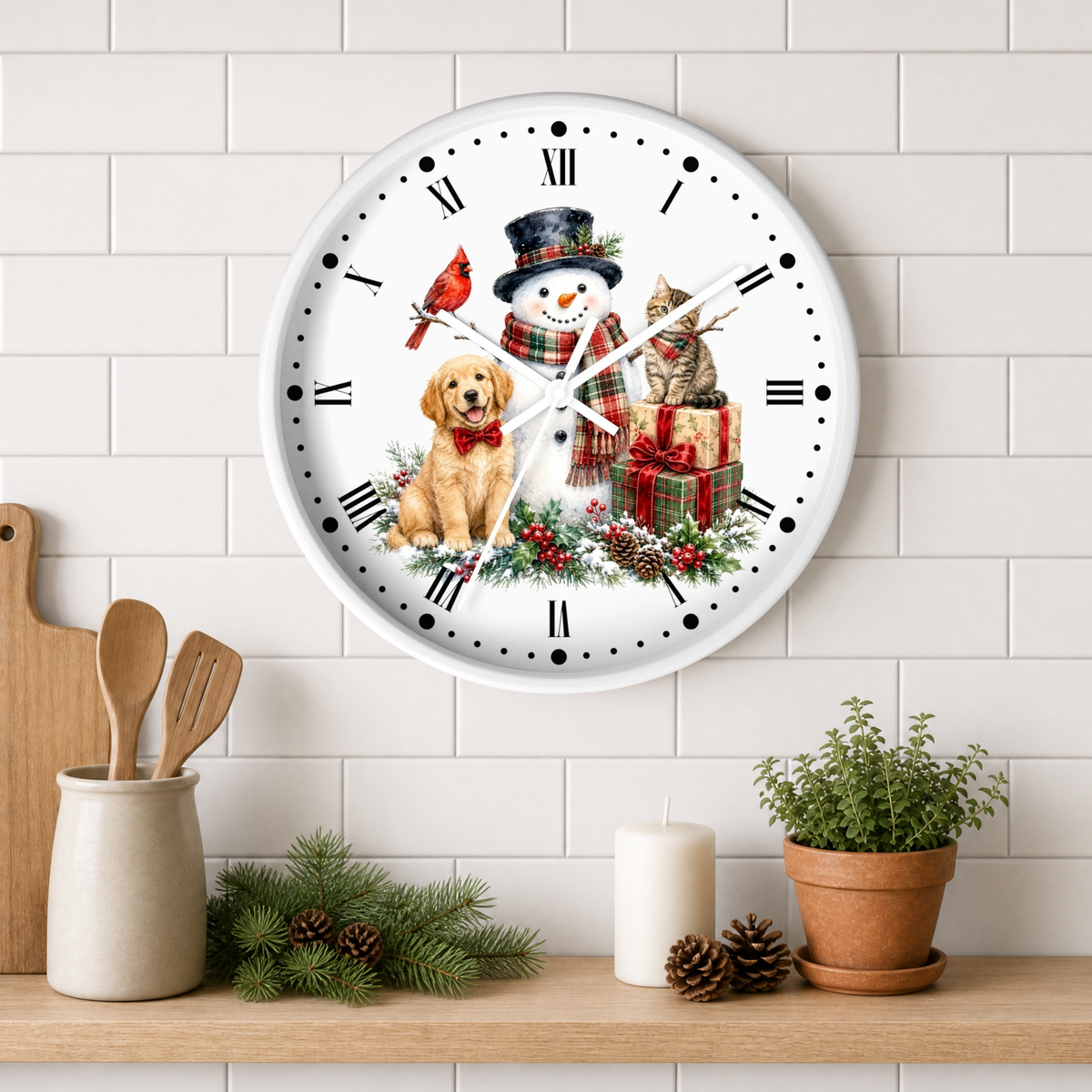 A round white wall clock with a festive Christmas design, featuring a snowman, golden retriever, tabby cat, gifts, and a cardinal. The clock is styled on a white subway tile wall above a wooden shelf with kitchen utensils, pine branches, a candle, and a potted plant.