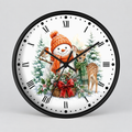 A round wall clock with a black frame and a white face featuring a festive Christmas illustration. The design includes a snowman in an orange knit hat and scarf, a robin, a deer, evergreen trees, and a basket with a red bow.