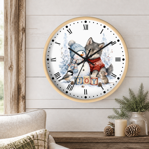 A round wall clock with a light wood frame hangs on a white shiplap wall above a wooden table. The clock face features a winter illustration of a gray wolf, a snowman in a blue knit hat and scarf, a small squirrel, a chickadee, and the word 'JOY' in colorful letters.