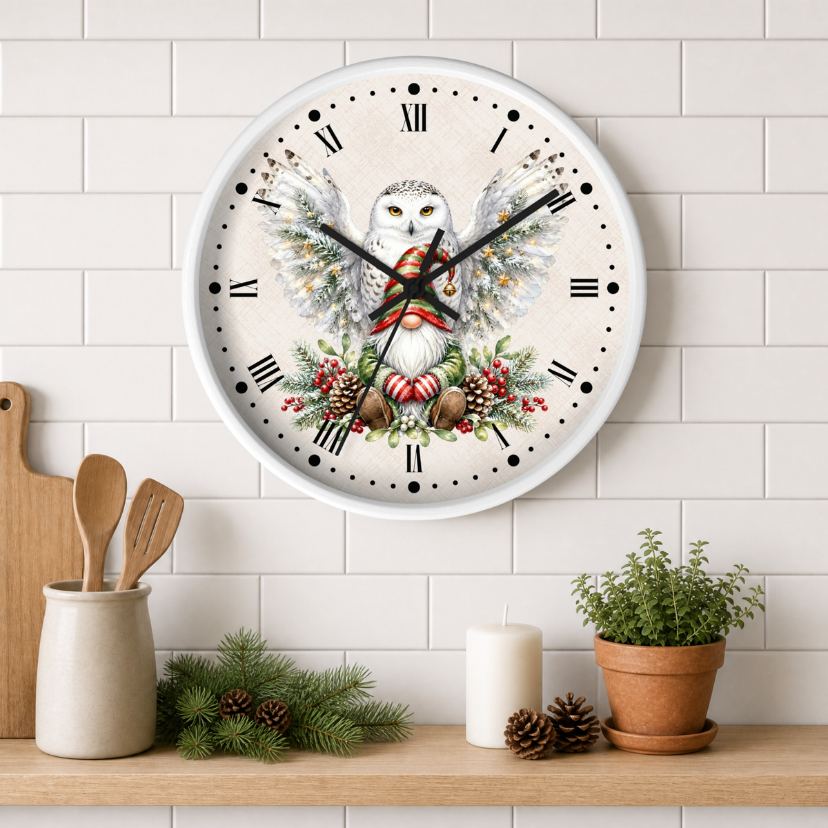 A round wall clock with a white frame and Roman numerals, featuring a detailed illustration of a white owl with spread wings holding a red ornament, surrounded by pinecones and greenery. The clock is mounted on a white subway tile wall above a wooden shelf styled with kitchen utensils, a candle, pinecones, and a potted plant.