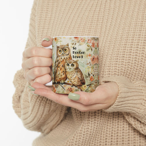 Person holding a mug with owl design and 'So Hooten Loved' text, wearing a beige sweater.