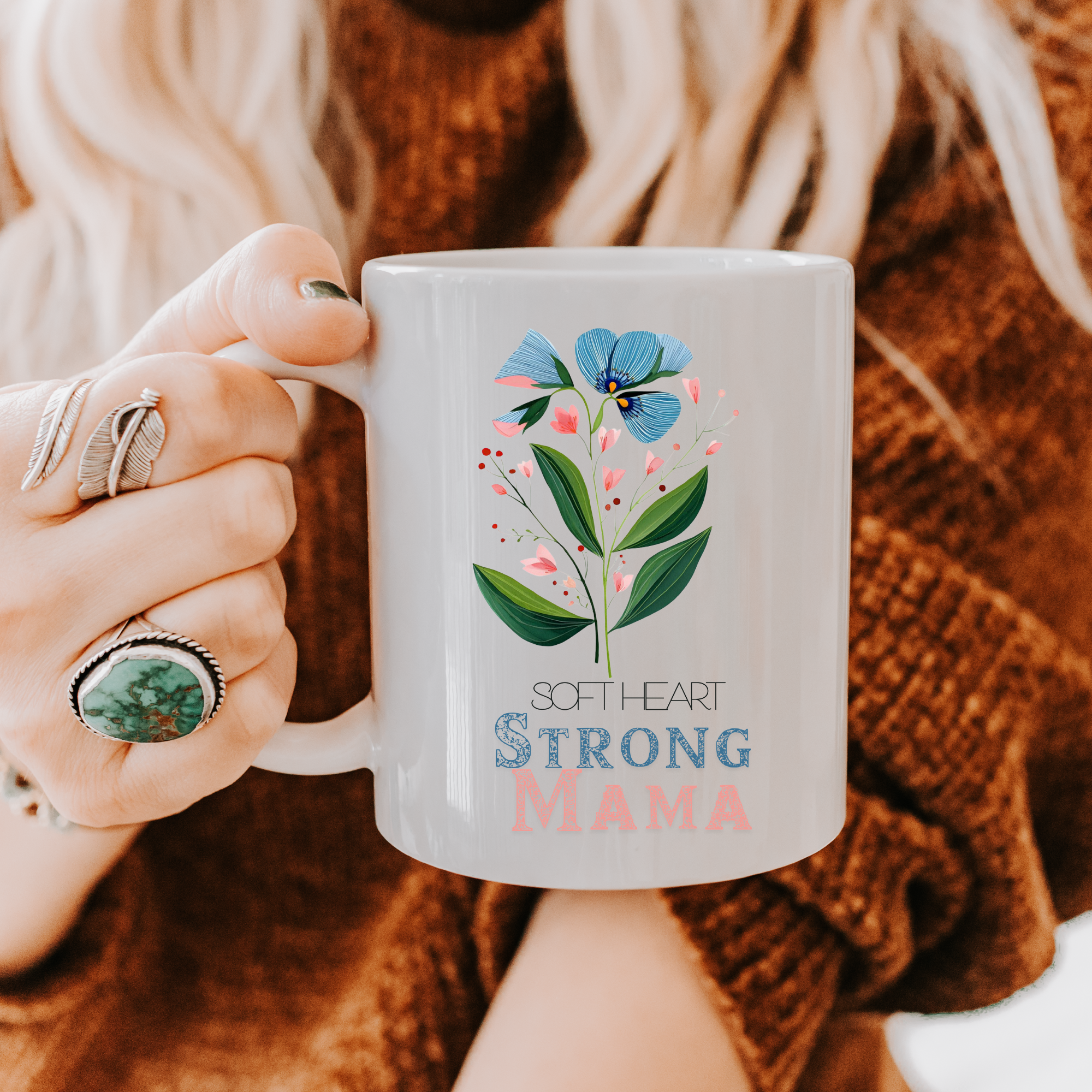 Person holding an 11oz white ceramic mug with floral design and text 'Soft Heart Strong Mama' against a blurred background.