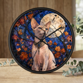 A round wall clock with a black frame featuring a stained-glass-style illustration of a hairless cat in front of autumn leaves and a full moon on a blue background, displayed on a wooden surface with eucalyptus branches.