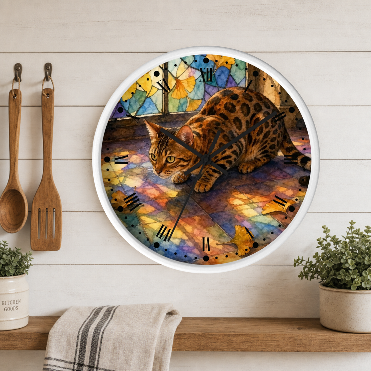 A round wall clock with a white frame hangs on a white wood plank kitchen wall above a wooden shelf. The clock face features a colorful stained-glass style background with a spotted brown cat walking across it.