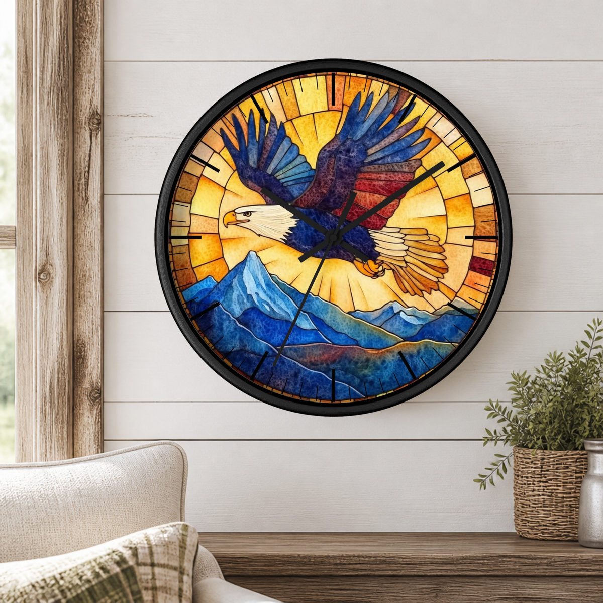 A round wall clock with a stained-glass-style design featuring a bald eagle flying over blue mountains, mounted on a white shiplap wall above a wooden table with a plant and part of a chair visible.