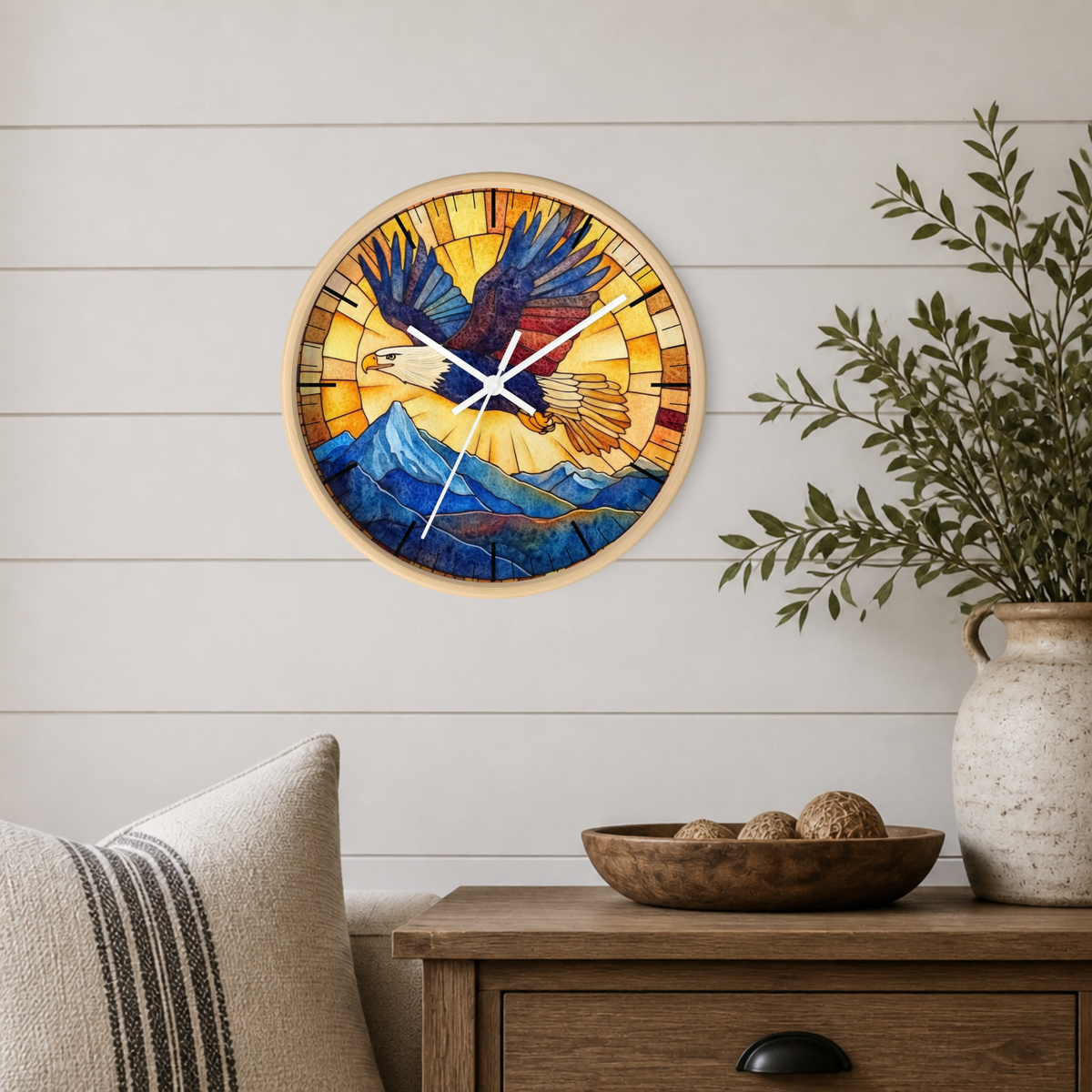 A round wall clock with a stained-glass-style eagle and mountain design, mounted on a white shiplap wall above a wooden side table with a vase and bowl.