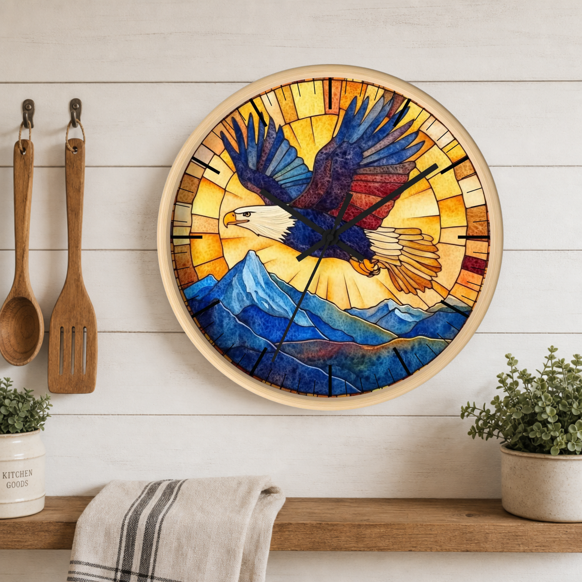 A round wall clock with a stained-glass-style design featuring a bald eagle flying over blue mountains, mounted on a white wood plank wall above a wooden shelf with plants and kitchen utensils.