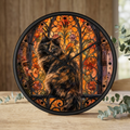 A round wall clock with a black frame and Roman numerals, featuring a fluffy calico cat sitting in front of an orange stained-glass style floral background. The clock is displayed on a wooden surface with eucalyptus leaves and a blurred indoor background.