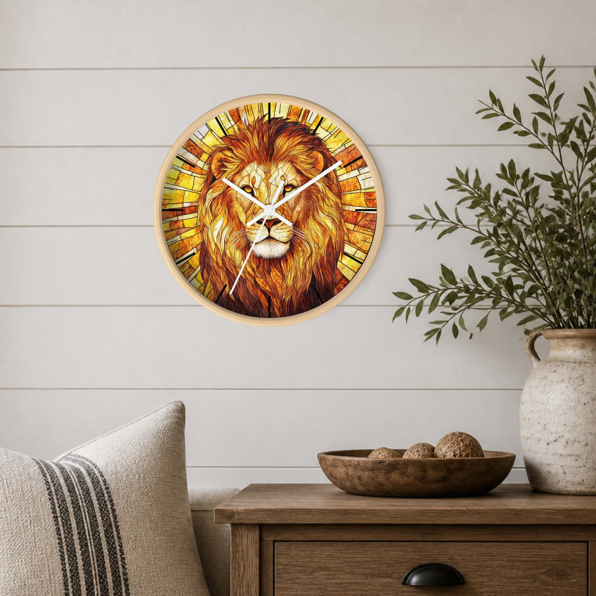 A round wall clock featuring a detailed lion illustration in warm golden, orange, and brown tones, mounted on a white shiplap wall above a wooden side table with a plant and decorative items.