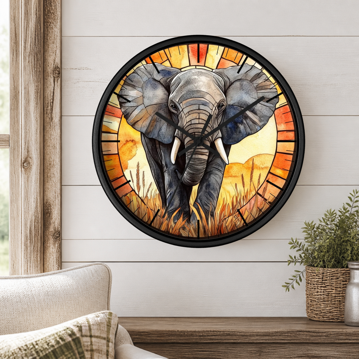 A round wall clock featuring a stained-glass-style illustration of an elephant in warm orange, yellow, and red tones, mounted on a white shiplap wall above a wooden shelf with a plant and part of a sofa visible.
