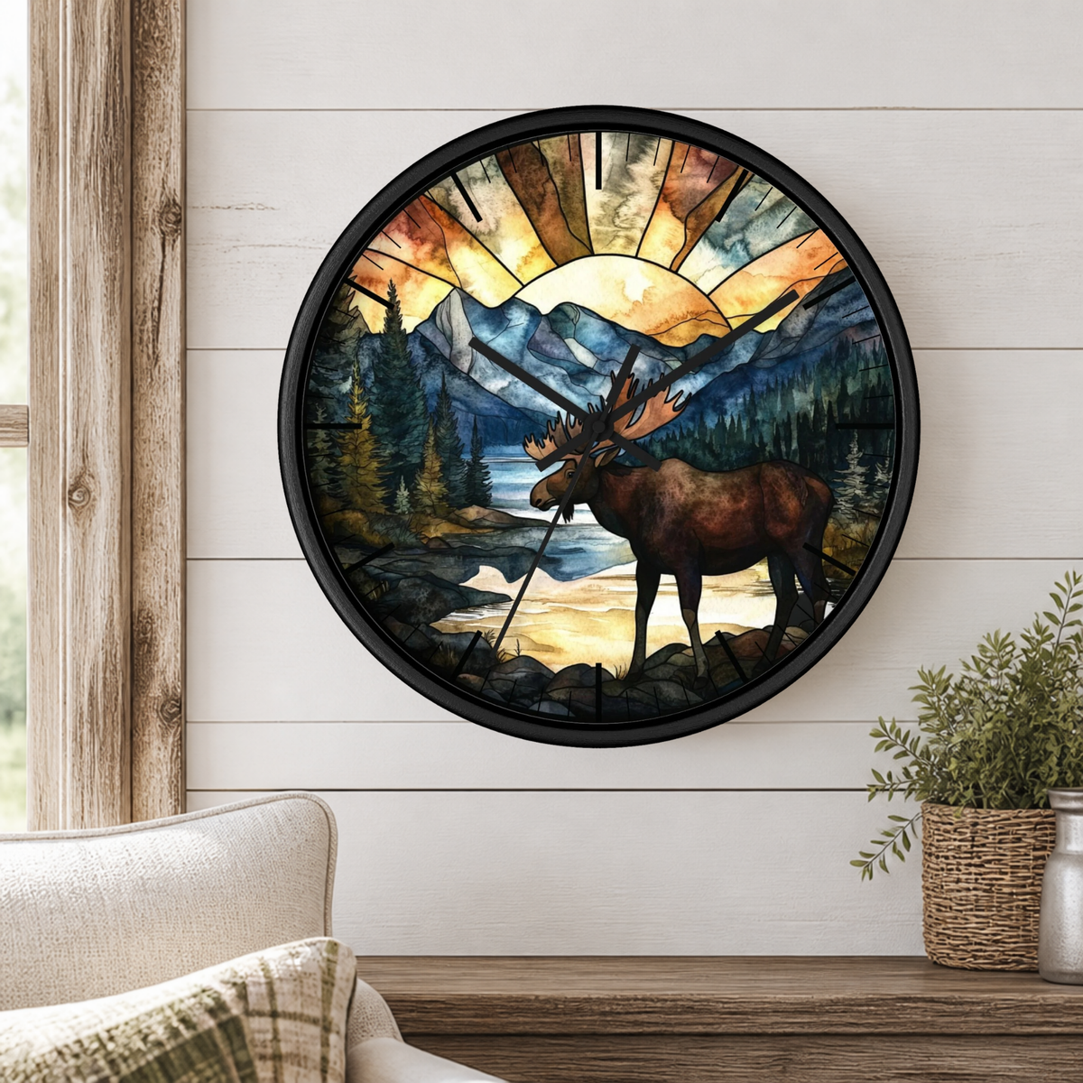 A round wall clock with a black frame and a stained-glass-style nature design featuring a moose, mountains, trees, and a sunburst, mounted on a white wood-paneled wall above a wooden shelf with a plant.