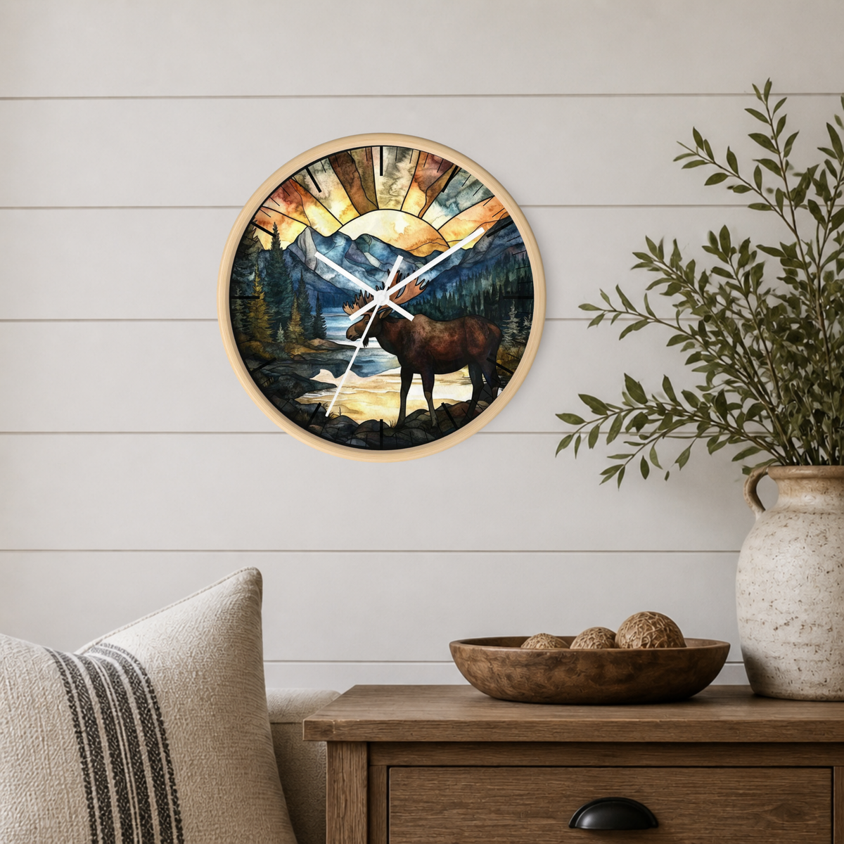 Round wall clock with a natural wood frame and a scenic moose landscape print, mounted on a white shiplap wall above a wooden side table with a vase and bowl.