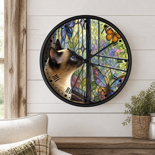 A round wall clock with a black frame and a stained-glass-style design featuring a Siamese cat, butterflies, and flowers, mounted on a white wood-paneled wall in a living room setting.