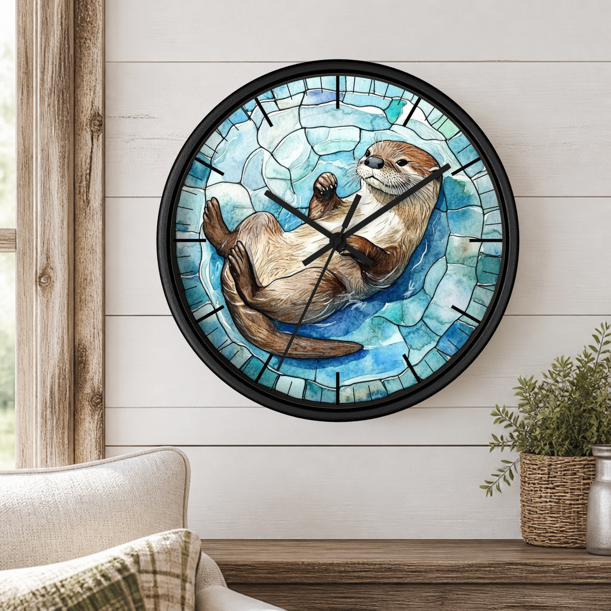 A round wall clock with a black frame and a stained-glass-style illustration of an otter on a blue icy background, mounted on a white shiplap wall above a wooden table with a plant and part of a chair visible.