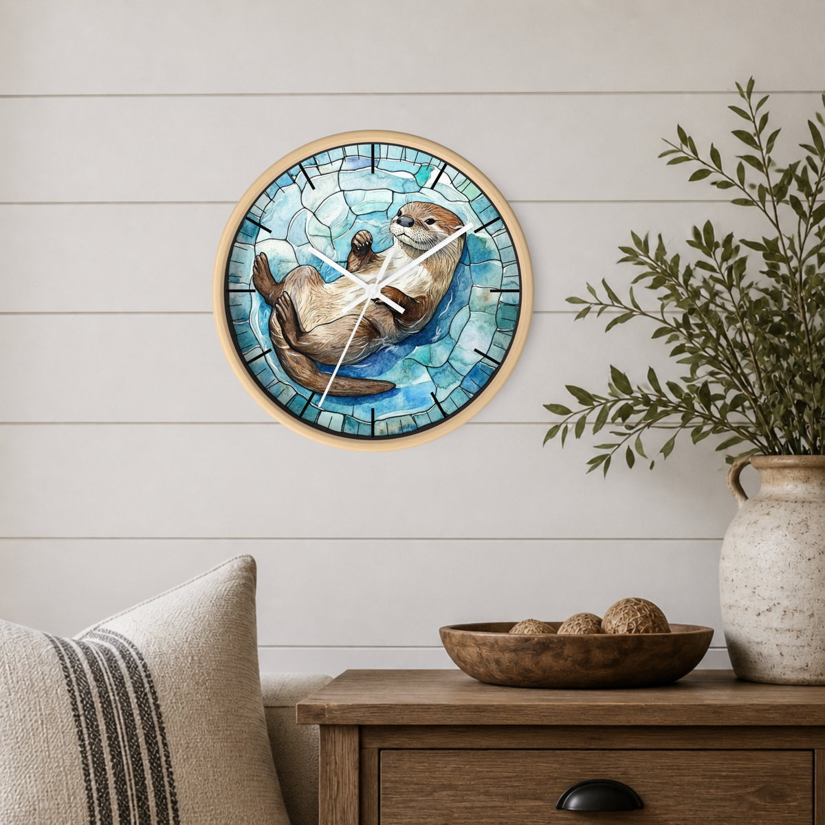 A round wall clock with a light wood frame and a stained-glass-style illustration of an otter on blue water, mounted on a white shiplap wall above a wooden side table with a vase and bowl.