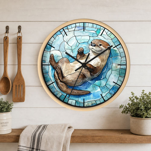 A round wall clock with a stained-glass-style design featuring a sleeping otter on blue and teal water, mounted on a white wood-paneled wall above a shelf with plants and kitchen utensils.