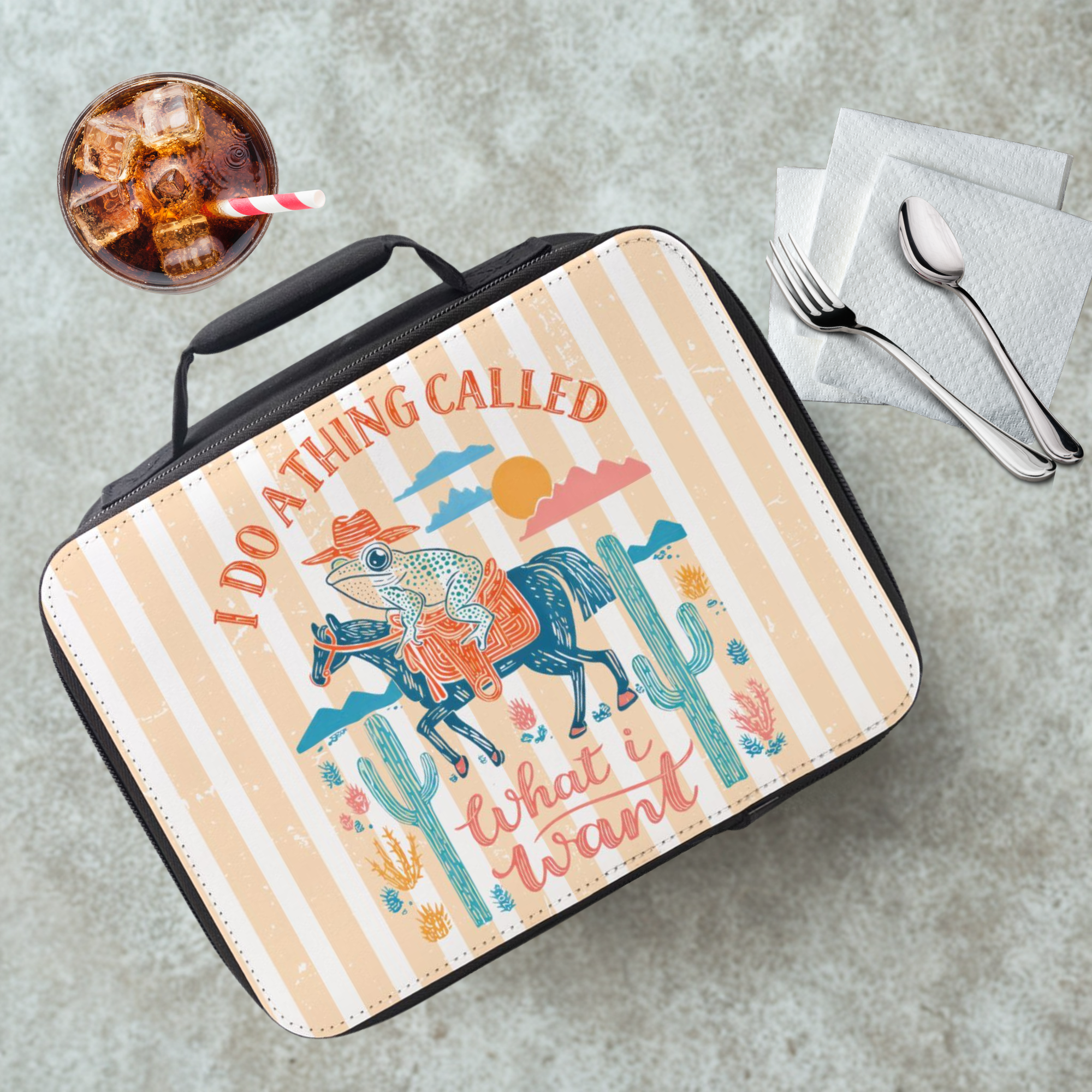  insulated lunch bag with a colorful striped design of a frog on a horse, cacti, and text on a gray table with a drink and utensils.