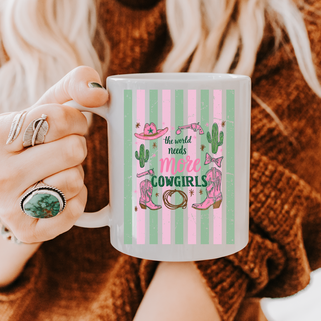 Person holding an 11oz mug with a 'more cowgirls' design against a textured brown background