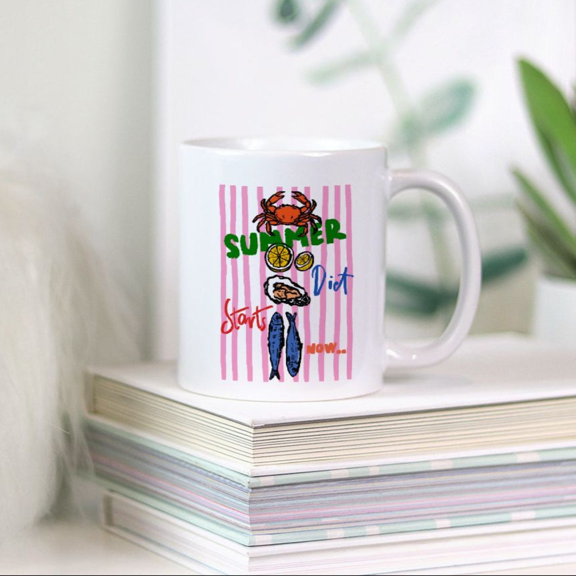 White 11oz. mug with colorful design on a stack of books with plants in the background