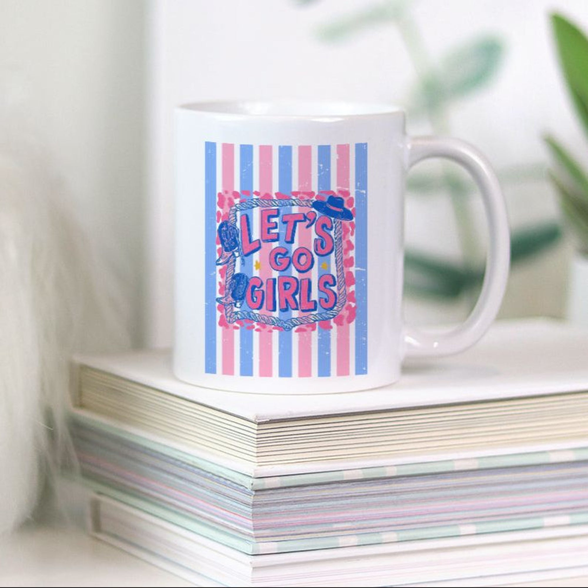 White 11oz. mug with colorful pink and blue striped design and text 'Let's Go Girls' on a stack of books with a plant in the background
