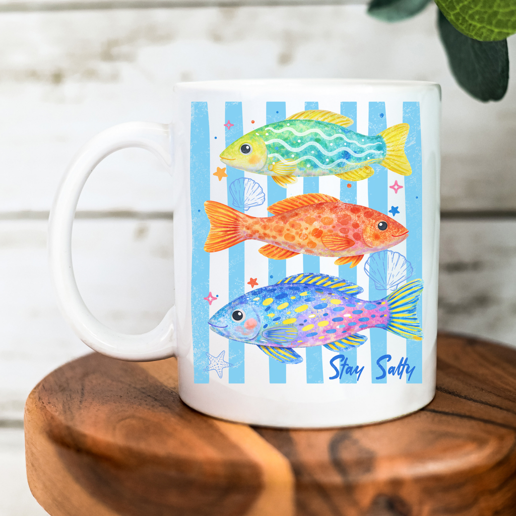 White mug with colorful fish illustrations and 'Stay Salty' text on a wooden surface.