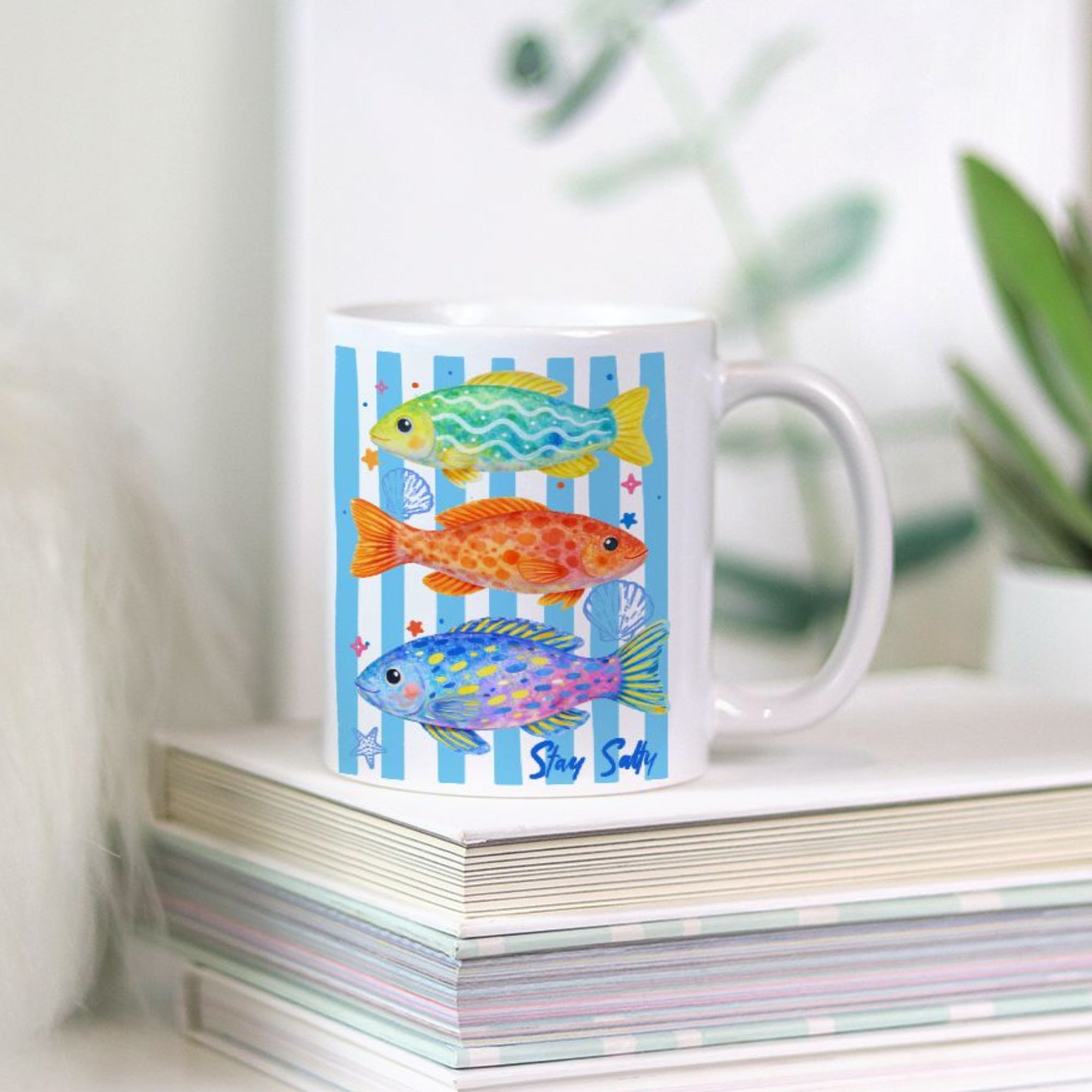Mug with colorful fish design on a stack of books with a plant in the background