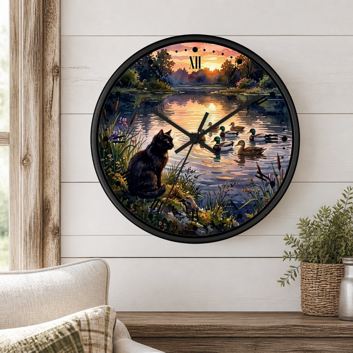 A round wall clock with a black frame displays a scenic print of a black cat sitting by a lake at sunset while ducks swim in the water. The clock is mounted on a white shiplap wall above a wooden mantel with a small potted plant.