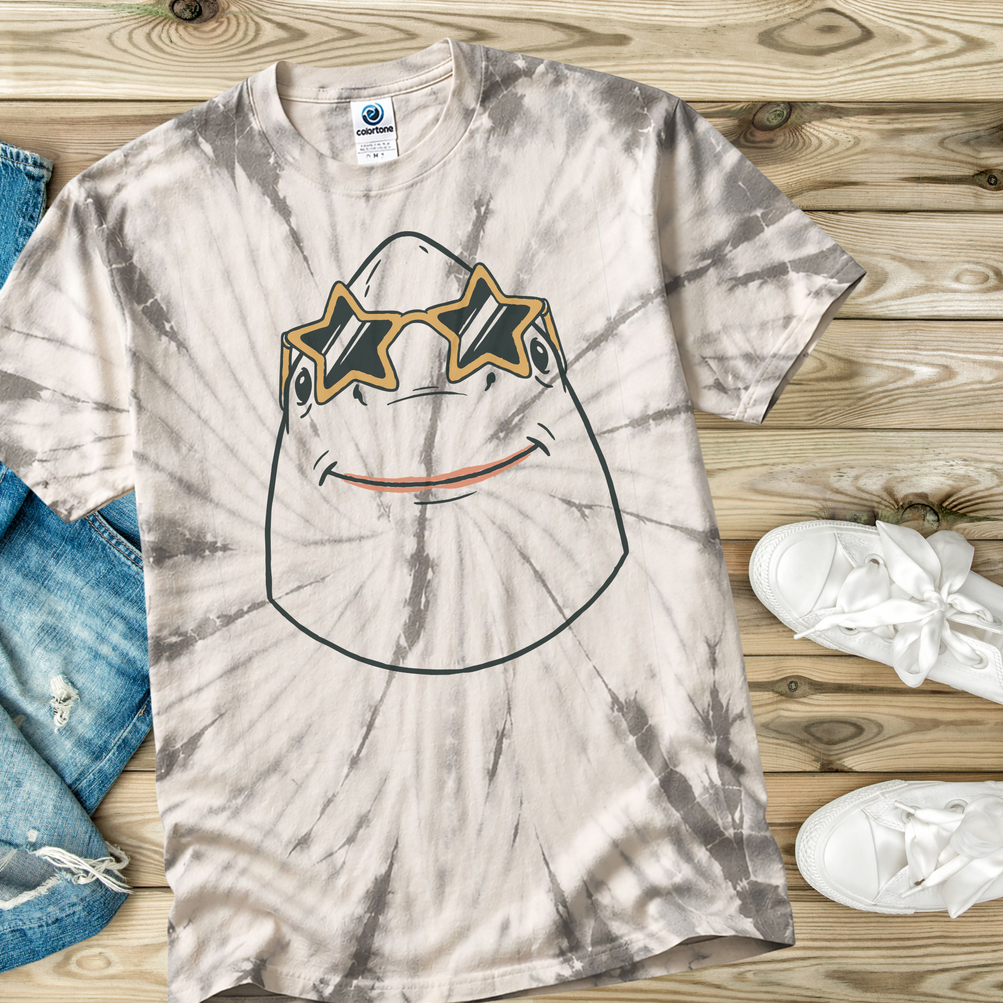 Tie-dye t-shirt  in the color Spider Silver with a smiley face shark wearing star shaped sunglasses design on a wooden surface with jeans and sneakers.