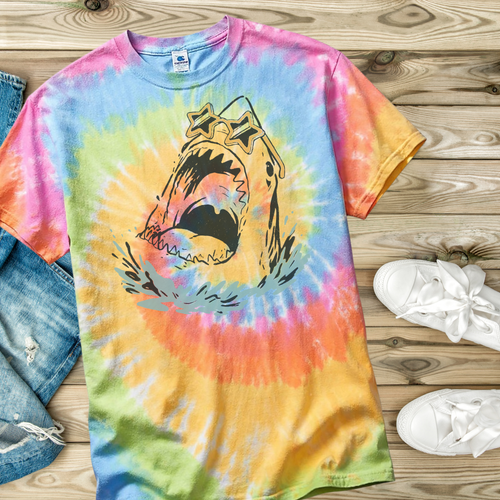 Tie-dye t-shirt with a hand drawn shark graphic on a wooden surface with jeans and shoes.