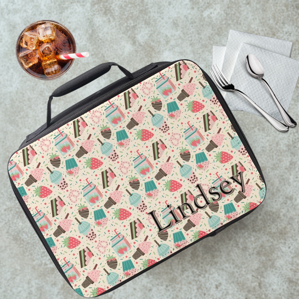 black lunch bag with a pattern of strawberries, milk, ice cream, cake, and personalized with a name on a cafeteria table with a drink and untensils.