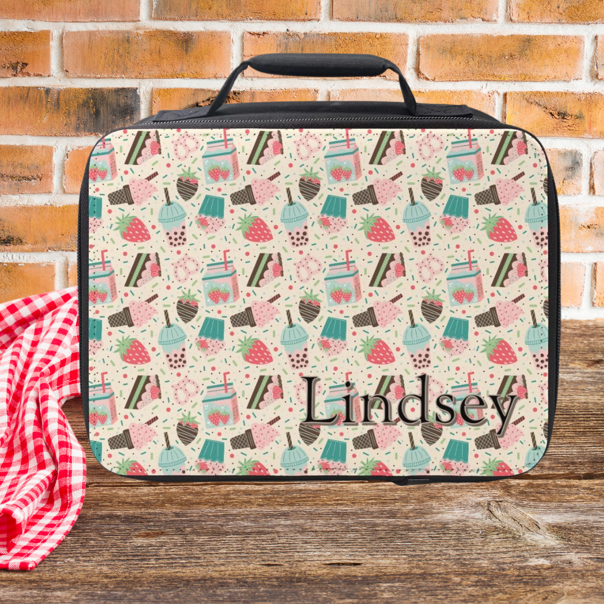 Black insulated lunch bag with a strawberry desert pattern personalized with a name on a wooden picnic table and brick wall background.