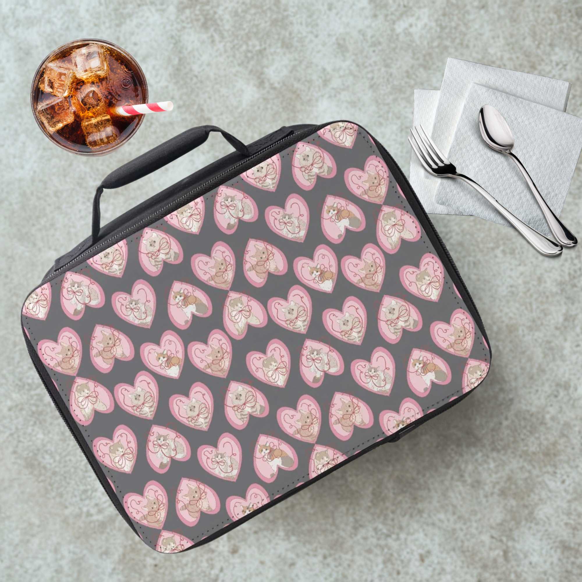 Lunch bag with hand drawn pink hearts and cats pattern on a gray table with a drink and utensils.