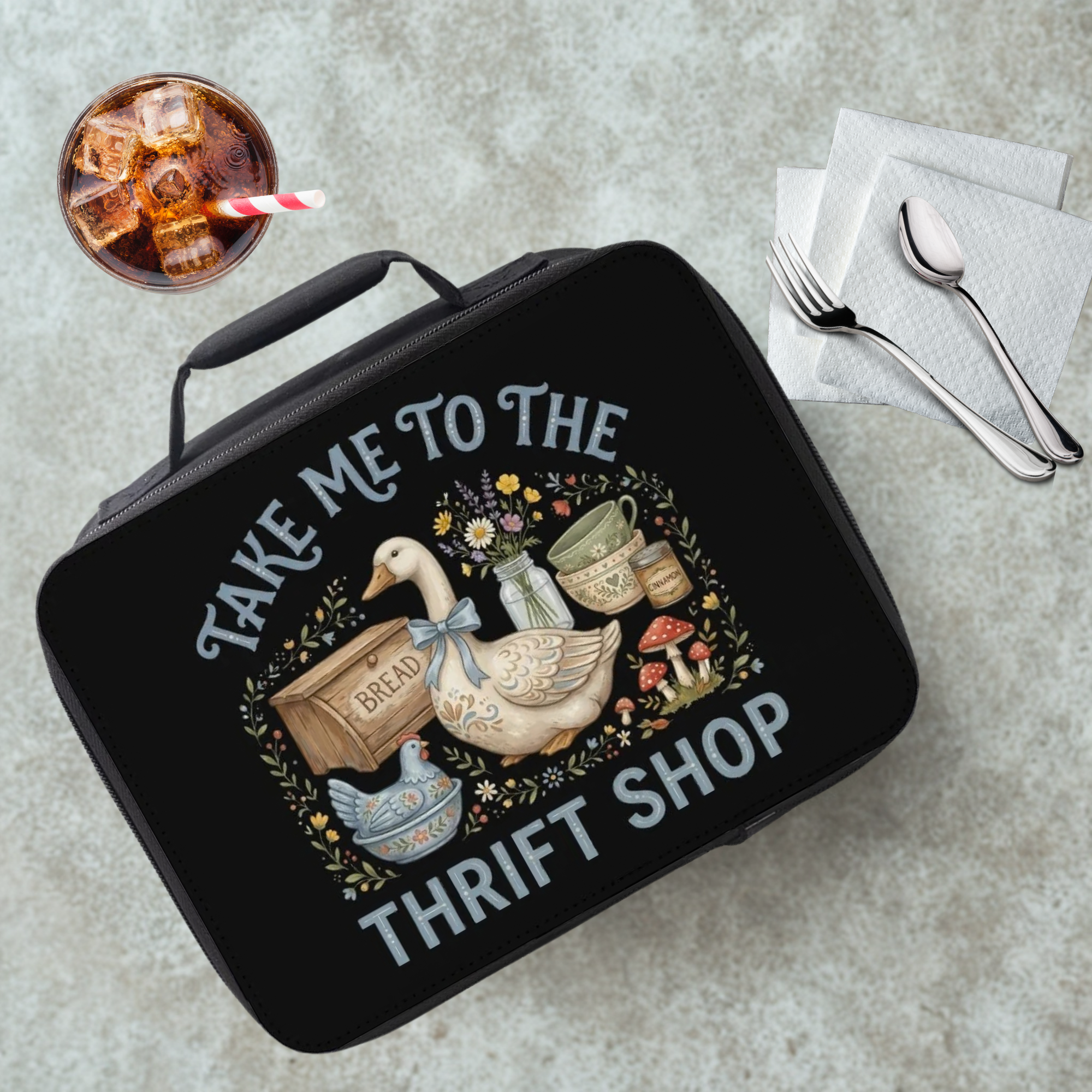 Black lunch bag with 'Take Me to the Thrift Shop' design on a cafeteria table with a drink and utensils.